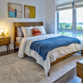 Beautiful bedrooms to fit every need 2-Bedroom Modern Apartments In Dorchester, MA - Imprint - Spacious Bedroom With Wood-Style Flooring, Wood-Frame Bed, Nightstand With Lamp, Desk, And Expansive Windows With Harbor View.